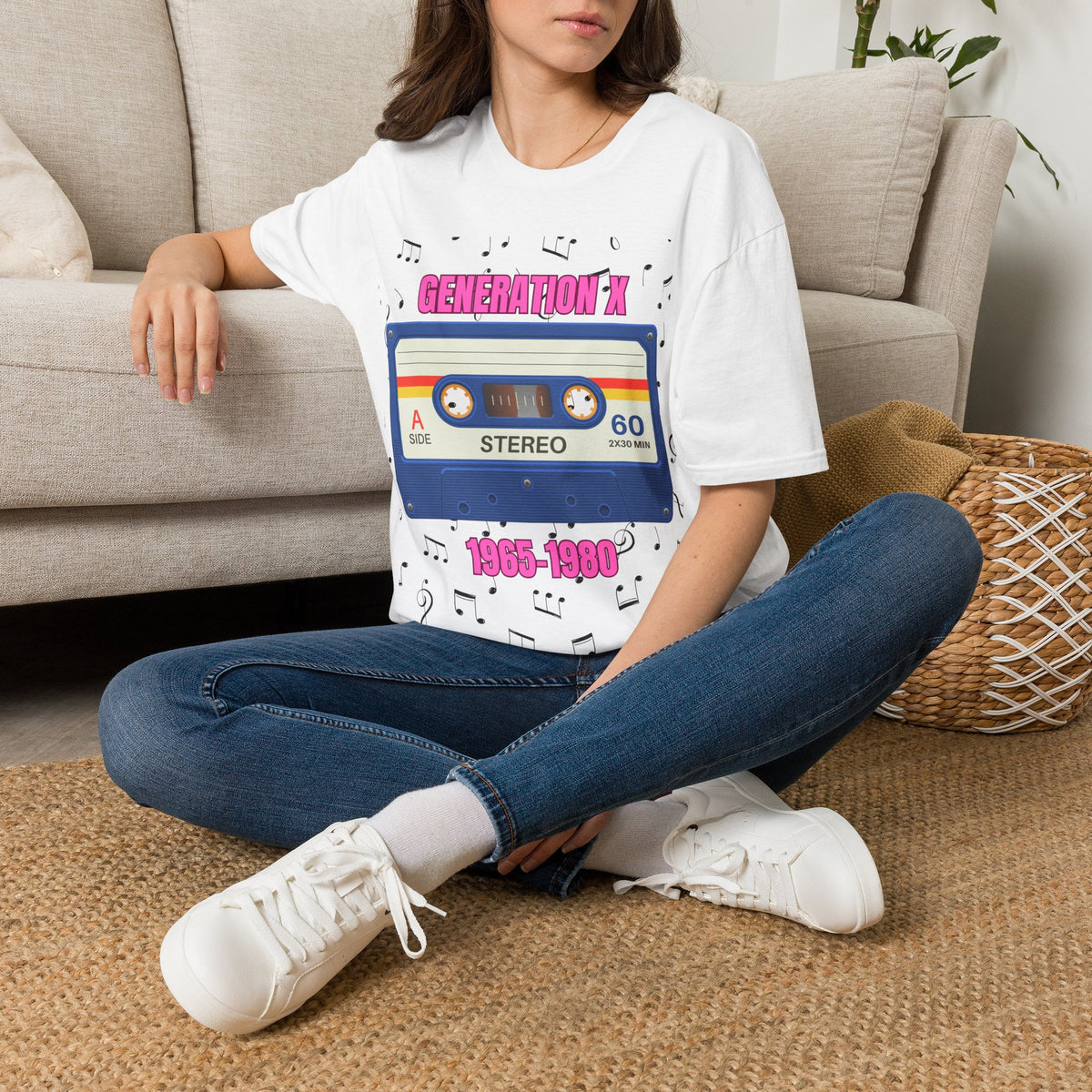 A woman sitting cross-legged on a woven rug in front of a beige sofa, wearing a white graphic T-shirt with a retro cassette tape design and pink text "GENERATION X 1965-1980" with blue jeans and white sneakers